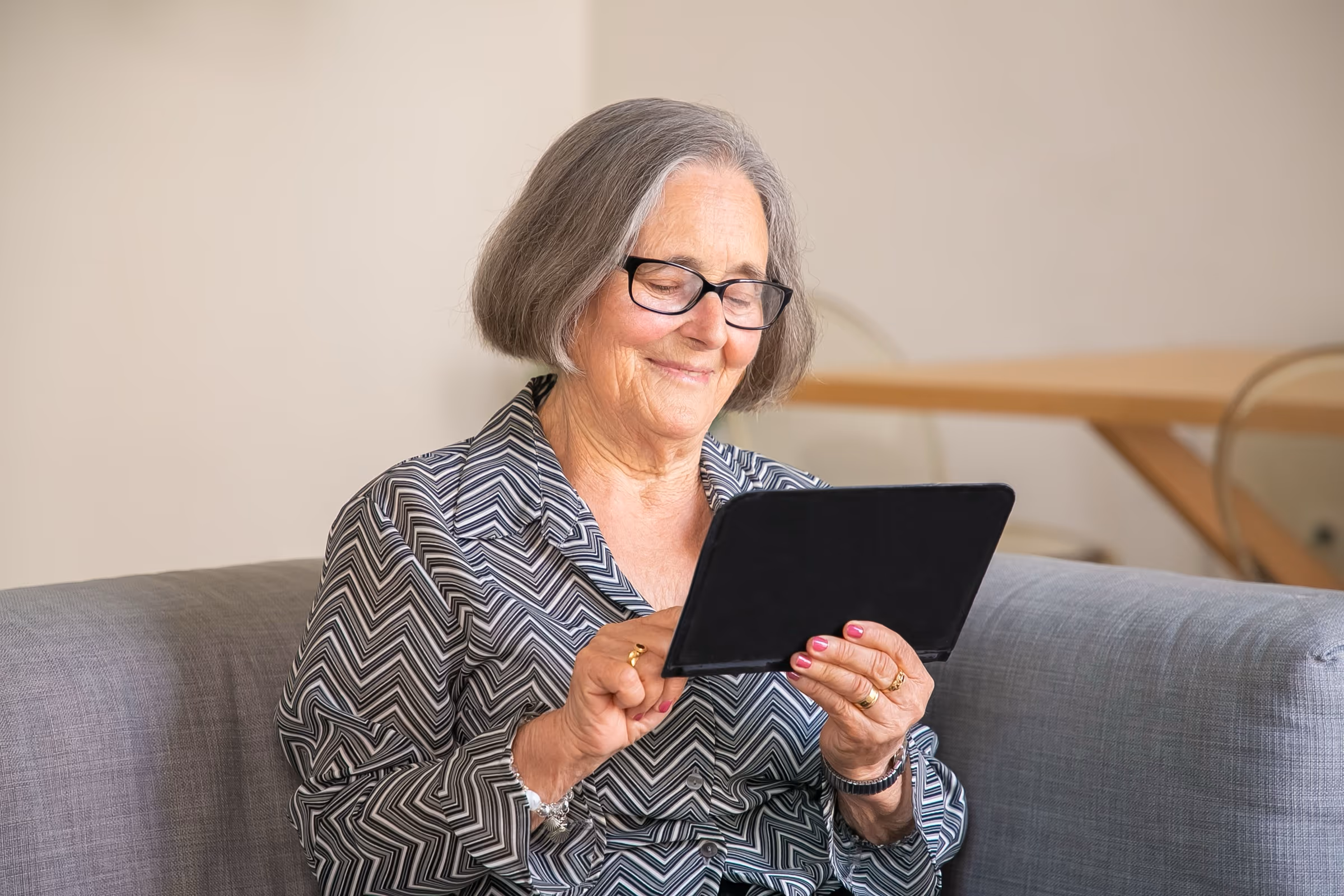 Ältere Frau mit Brille, die auf dem Sofa lächelnd auf ihr Tablet schaut
