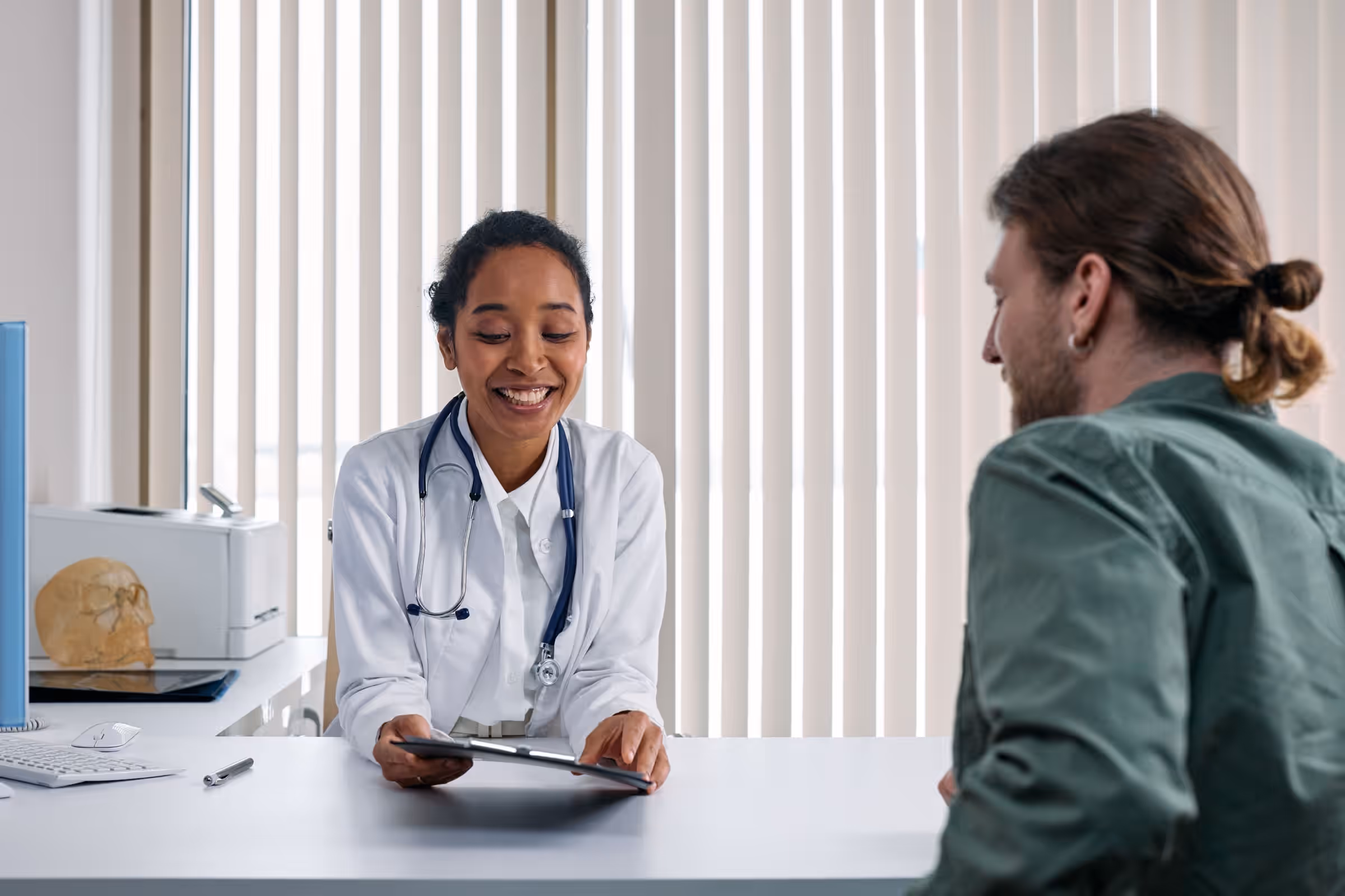 Lächelnde Ärztin im Kittel am Schreibtisch im Gespräch mit einem Patienten
