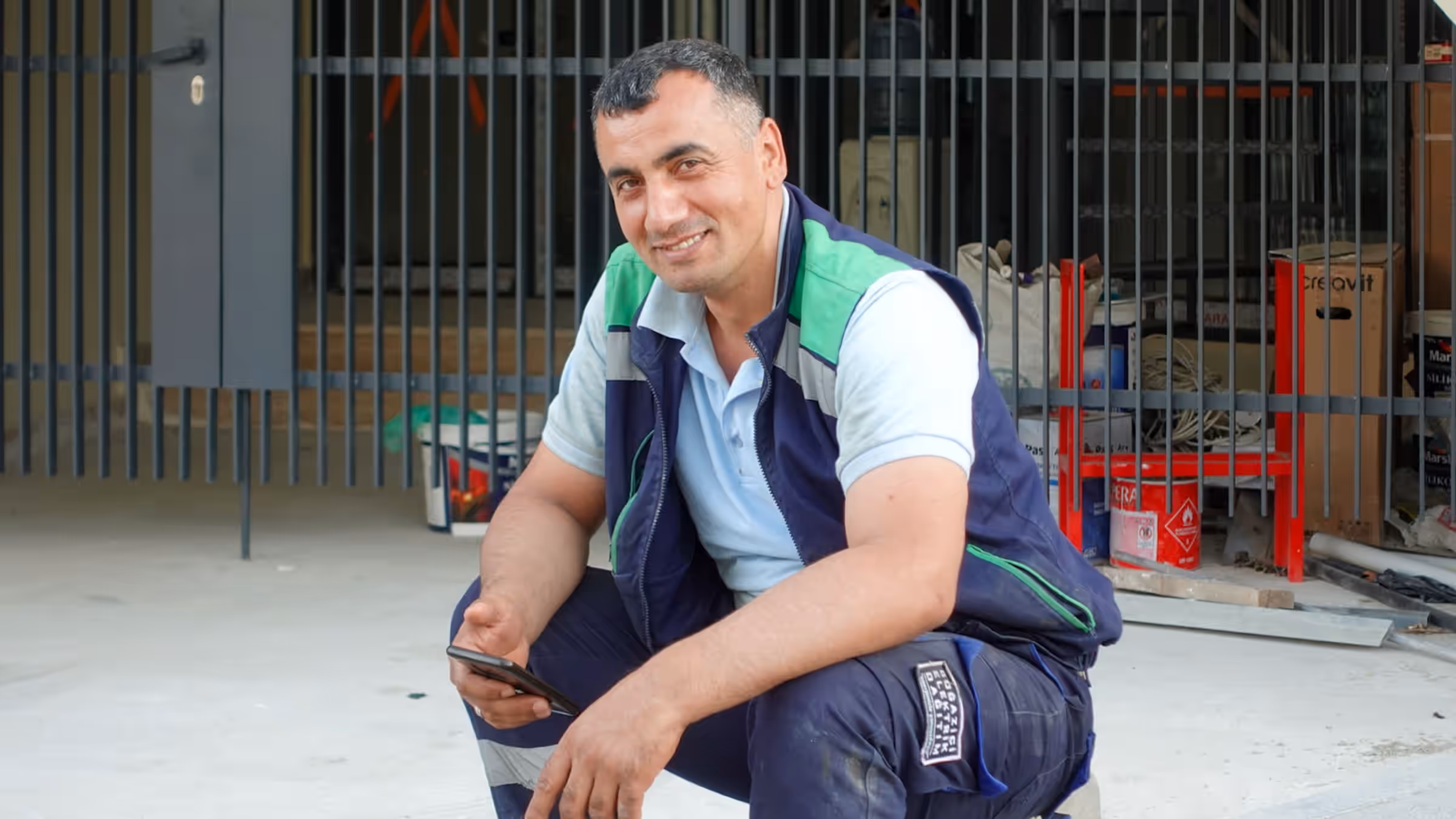Handwerker in Arbeitskleidung vor einer Baustelle, der lächelnd von seinem Handy aufschaut
